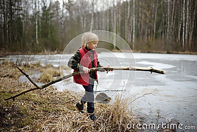 Little boy playing large branch on shore of forest lake on early spring day. Surface of lake is still under ice. Child tries to Stock Photo