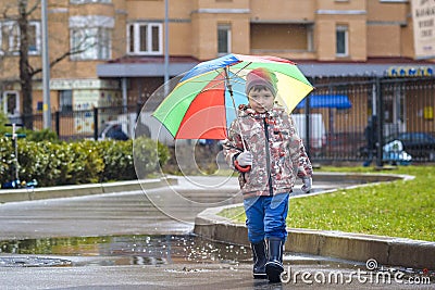 Little boy playing in rainy summer park. Child with colorful rainbow umbrella, waterproof coat and boots jumping in puddle and mud Stock Photo
