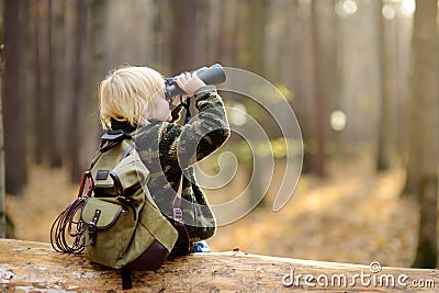 Little boy scout with binoculars during hiking in autumn forest. Child is sitting on large fallen tree and looking through a Stock Photo