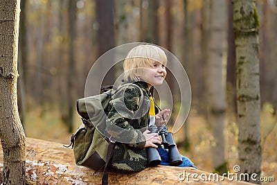 Little boy scout with binoculars during hiking in autumn forest. Child is sitting on large fallen tree and looking through a Stock Photo