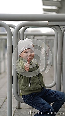 Little boy sits on an iron fence in a park and shows thumb Stock Photo