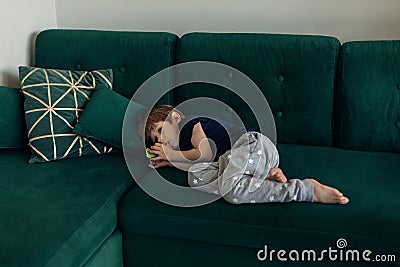 Little boy in sleepwear lying on big green modern sofa in living room and sucking finger. Bad habbit and manners of Stock Photo