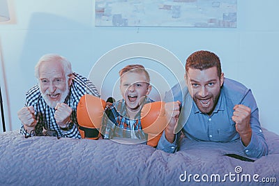 Little boy wearing boxing gloves, his father and grandfather lying on the bed, watching a boxing match Stock Photo