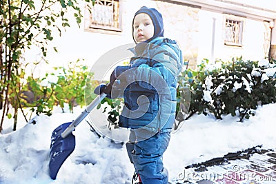 Little caucasian boy shovels snow in the yard with beautiful snowy rose bushes. Child with shovel plays outdoors in winter. Childr Stock Photo