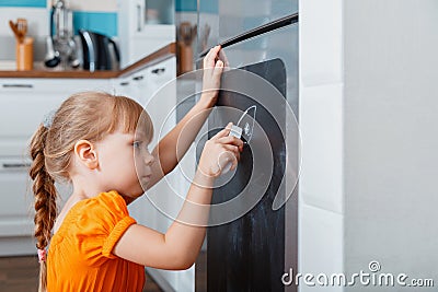 Little caucasian focused child girl draw with chalk on chalk board on refrigerator in kitchen at cozy home interior. Stock Photo