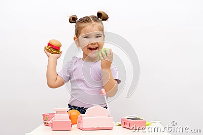 Little caucasian smiling girl with toy hamburger and apple in hand is playing in store, toy cash register, vegetables, basket, Stock Photo