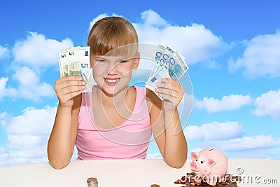 Little lovely girl sitting on the desk with coins and pink piggy bank and holding in hands european currency over blue sky Stock Photo