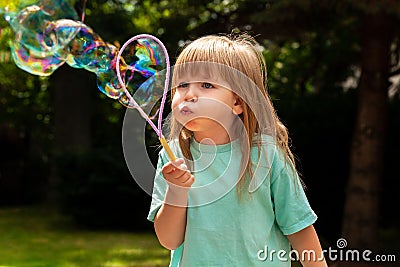 Little child, girl blowing huge bubbles alone, portrait outdoors. Young kid with puffed cheeks blows big bubbles outside, closeup Stock Photo