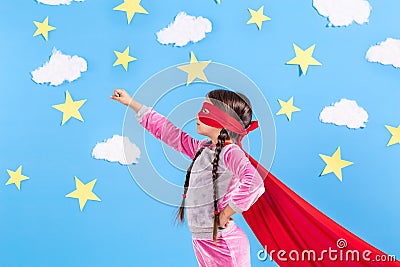 Little child plays superhero. Kid on the background of bright blue wall with white clouds and stars . Girl power concept Stock Photo