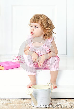 Little curly girl in a pink dress sits with a thoughtful look and watch into the distance, on a white porch. Stock Photo