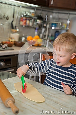 Little cute blond boy in kitchen is learning to cut flour dough with children`s shaped baking knife. Concept of family leasure Stock Photo