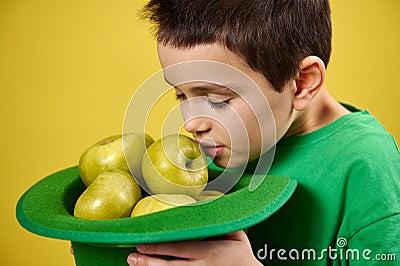 Little cute boy enjoys the scent of green apples in a green Irish leprechaun cap. Face portrait on yellow background. Saint Stock Photo