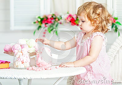 Little cute curly girl in a pink dress with lace and polka dots sitting at the table and eating different sweets. Stock Photo