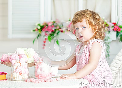 Little cute curly girl in a pink dress with lace and polka dots sitting at the table and eating different sweets. Stock Photo