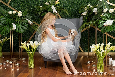 Little cute girl with blond hair in a white dress holding a small dog and white flowers, lilies and orchids Stock Photo