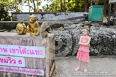 Little cute girl standing near monkey golden statuettes and macaco at zoo in Thailand. Stock Photo