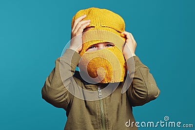 Cute kid looking at camera thru yellow knitted thief mask, over blue Stock Photo