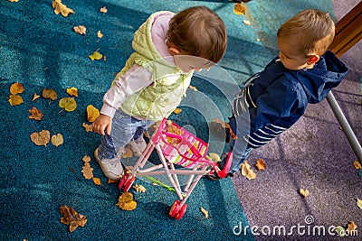 Little cute toddler boy and a beautiful year-old baby girl in blue clothes play in a pink toy stroller for a doll in the Stock Photo