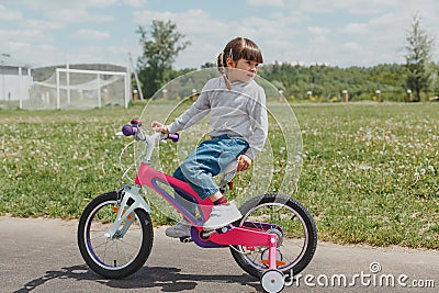 Little cute todler girl in jeans and a sweatshirt with pigtails sits on a pink bike at the stadium in summer, does not look at the Stock Photo