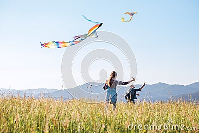 Little daughter with the father running fast while they flying up colorful kites on the high grass meadow in the mountain fields. Stock Photo