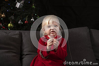 Little disheveled girl in a red dress sits on the sofa against the background of a Christmas tree. Christmas and new year`s eve Stock Photo