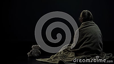 Little female kid in plaid sitting near teddy bear in dark room, loneliness Stock Photo