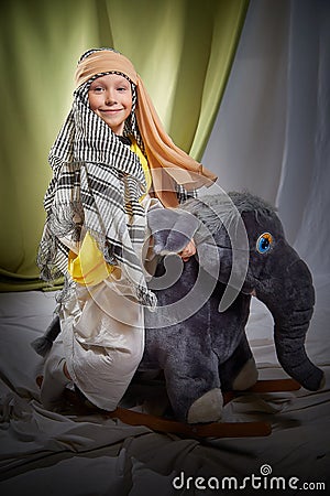 Little funny boy in long white and yellow dress with scarf on head and elephant toy. Young male model posing for photo Stock Photo