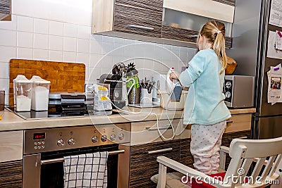 Little girl baking waffles in the kitchen following a recipe on the smartphone while standing on a chair Stock Photo