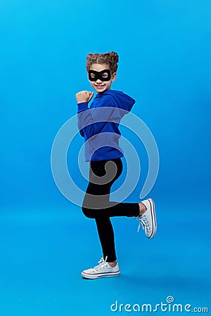 Little girl in black mask, plays, cute robber smiling. Child masked bandit, over blue background. Vertical view. Stock Photo