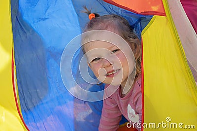 A little girl with blond curly hair looks out from the multicolored tent and smiles Stock Photo