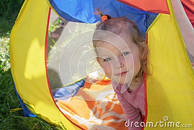 A little girl with blond curly hair looks out from the multicolored tent and smiles Stock Photo