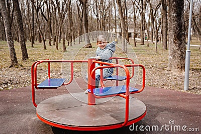 A little girl in a blue coat with a plush toy spins on carousel on playground. Stock Photo