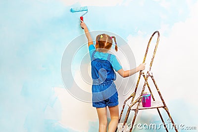 Little girl in blue paints the wall on a ladder Stock Photo