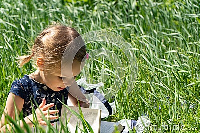 Little girl with a book in the garden. Kid is readding a book. A little girl 4-5 years old sits on the grass and reads a book Stock Photo