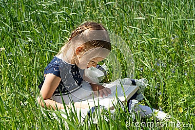 Little girl with a book in the garden. Kid is readding a book. A little girl 4-5 years old sits on the grass and reads a book Stock Photo