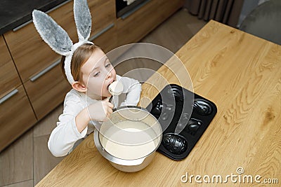 Little girl in bunny ears eating, licking sweet dough for muffins or cake with spoon from bowl in modern kitchen Stock Photo