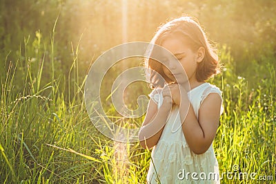 Little Girl closed her eyes, praying in a field during beautiful sunset. Hands folded in prayer concept for faith Stock Photo