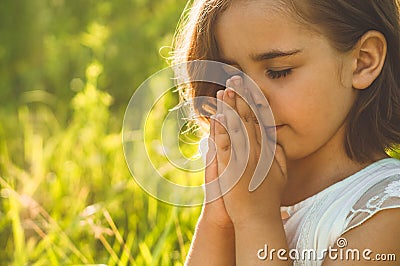 Little Girl closed her eyes, praying in a field during beautiful sunset. Hands folded in prayer concept for faith Stock Photo