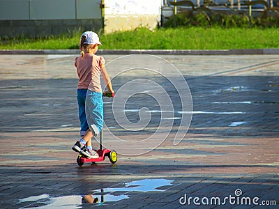 A little girl in colorful summer clothes is riding a children`s scooter among puddles Stock Photo