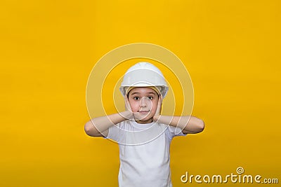 Little girl in a construction white helmet covers her ears from loud sounds on a yellow background Stock Photo