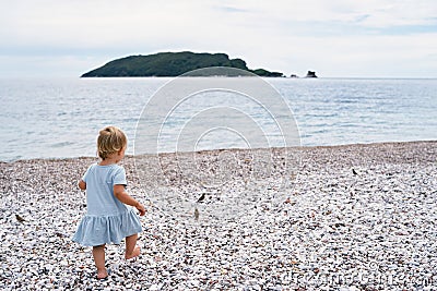 Little girl in a dress walks along a pebble beach to the sea with an island in the distance Stock Photo