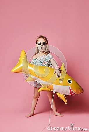 Girl wears a balloon in the shape of a yellow shark-fish, celebrates a holiday, smiles broadly, stands against a pink background Stock Photo