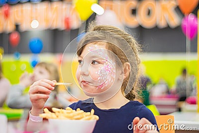 A little girl eats french fries in a restaurant. A child in a beautiful makeup eats in the nursery Stock Photo