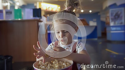 Little girl eats popcorn in movie theater. Media. Cute girl eating popcorn in waiting room at cinema. Pretty girl cute Stock Photo