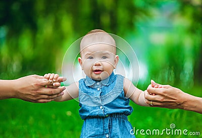 The little girl with a fair, short hair in a jeans dress without sleeves takes the first steps in park in warm, summer day, parent Stock Photo