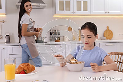 Little girl having breakfast while mother cooking food in kitchen. Single parenting Stock Photo