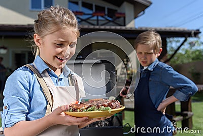Little girl holding plate with steaks while her brother preparing meat on grill Stock Photo