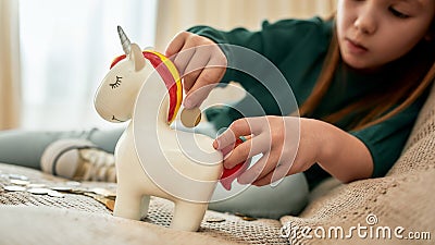A little girl holding a red tail of her porcelian unicorn moneybox and putting a coin into a slot while sitting on a Stock Photo