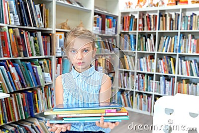 Little girl holds stack of books with fairy tales in children`s library. Special reading kids room. Shelves with bright Stock Photo