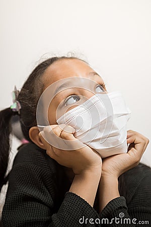 Little girl with medical mask looking up hoping that everything will turn out well. Girl rests her head on her hands during the Stock Photo